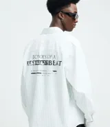 Camisa de manga larga con diseño de rayas verticales, cuello clásico y cierre frontal con botones. Confeccionada en una mezcla de algodón, poliamida y elastano, presenta una estampa gráfica en la espalda con la frase Echoes of a Restless Beat.