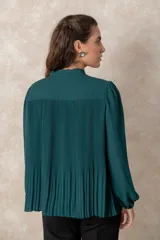 Blusa plisada de color verde oscuro, con mangas largas terminadas en puños elásticos y cuello alto con lazo frontal para anudar.