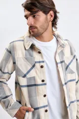 Sobrecamisa de tejido tipo polar con estampado de cuadros en tonos beige, gris y azul. Presenta cuello clásico, cierre frontal con botones oscuros y bolsillos frontales en el pecho.