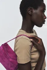 Mini bolso de mano confeccionado en piel de gamuza color fucsia. Presenta un diseño estructurado con asa corta, cierre principal mediante cremallera y solapa, y bolsillos exteriores en la parte frontal y trasera.