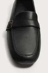 Mocasines de piel color negro, con punta redonda, suela ultrafina y detalle de tira con hebilla metálica dorada en el empeine.