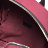 Mochila de diseño urbano en color bordó, confeccionada en material textil. Cuenta con compartimento principal con cierre, bolsillo frontal con cierre, manija superior y detalle de llavero trenzado en blanco y negro. Incluye logo circular de la marca en la parte inferior.
