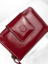 Mini billetera de cuero charolado color rojo, con cierre perimetral y solapa frontal con broche, decorada con un logo de búho grabado.