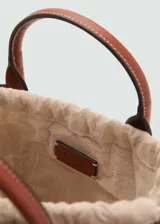 Bolso tipo cesta de fibras naturales con diseño en forma de abanico. Presenta detalles en cuero sintético marrón en el borde superior, asas cortas de mano y una correa bandolera larga ajustable. Incluye cierre de botón magnético y forro interior.