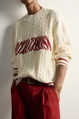 Jersey relaxed fit de punto tejido en hilatura de algodón, color crudo con detalles en bordó. Presenta cuello redondo, manga larga, diseño de trenzas y texto en intarsia a contraste en el delantero, con acabados en rib y líneas en los puños y el ruedo.