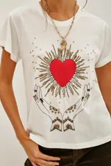 Remera blanca de algodón con estampado frontal de manos en posición de rezo rodeando un corazón rojo con rayos dorados.