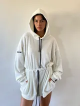 Vestido tipo hoodie oversize de tejido waffle, color crudo, con capucha y cordones ajustables en contraste. Presenta un cordón interno en la cintura para entallar la prenda y bolsillos laterales.