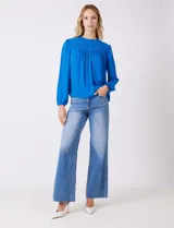 Blusa azul de gasa con pliegues en la parte delantera y detalles de encaje calado. Cuello alto fruncido y mangas largas con puños elastizados. Espalda lisa con abertura tipo gota y botón.