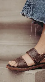 Sandalias planas de cuero color marrón chocolate, con dos tiras anchas en el empeine decoradas con costuras artesanales y tira ajustable con hebilla en el talón.