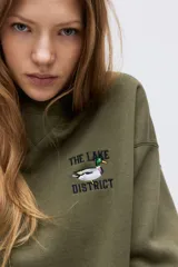 Buzo oversized color verde militar con interior cepillado suave. Presenta hombros caídos y un diseño de texto bordado en el pecho que dice "THE LAKE DISTRICT" con un pato. Tiene ribete acanalado en cuello, puños y bajo.
