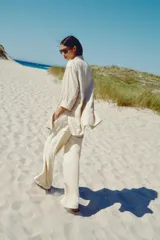 Pantalón ancho de tiro alto color beige claro, con cintura elástica, cordón ajustable y pinzas delanteras. Presenta bolsillos frontales y bolsillos de plastrón en la espalda. Incluye un cinturón del mismo tejido con apliques metálicos.