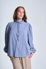 Camisa de color azul con cuello alto decorado con volados y lazo de ajuste. Presenta tablas decorativas en los hombros, cierre frontal con botones de nácar y mangas largas con puños elásticos que generan un efecto abullonado.