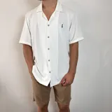 Camisa de manga corta tejida en color negro, con cuello tipo camp y cierre frontal de botones blancos. Presenta un calce holgado y un pequeño logo bordado en el pecho.