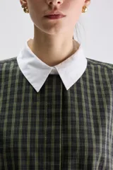 Vestido camisero midi de calce relajado a cuadros en tonos verde y negro, confeccionado en algodón liviano. Presenta cuello clásico en contraste de color blanco, cartera frontal oculta con botones y mangas cortas con vuelta.
