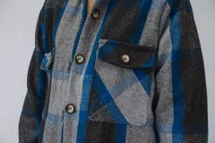 Tapado largo estilo camisa confeccionado en paño italiano, con diseño a cuadros en tonos de azul, gris y negro. Cuenta con cuello clásico, cierre frontal con botones, bolsillos con solapa en el pecho y mangas largas.