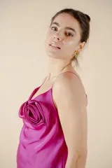 Vestido midi fucsia de satén, con escote buche, breteles finos y detalle de rosa de tela aplicada en el escote. Presenta un tajo lateral en la falda y calce al cuerpo.