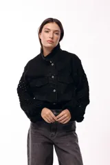 Campera de jean negra con mangas largas tejidas en crochet con diseño calado y detalles florales en relieve. Presenta cuello camisero, cierre frontal y bolsillos en el pecho.