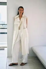 Chaleco largo fluido confeccionado en mezcla de lyocell, color crudo, con cuello de solapa, manga sisa, bolsillos frontales de plastrón y cinturón ajustable del mismo tejido.