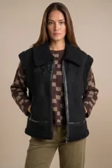 Chaleco estilo gamulán de color negro, con interior y detalles de corderito sintético. Presenta cierre frontal, cuello solapa y bolsillos laterales.