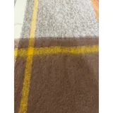 Bufanda rectangular de tejido grueso con diseño de cuadros en tonos azul, naranja, gris y beige, terminada con flecos en los extremos.