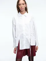 Camisa blanca de algodón con cuello clásico, cierre frontal de botones y diseño de paneles verticales. Presenta tiras largas irregulares en el frente que aportan movimiento y un estilo contemporáneo.