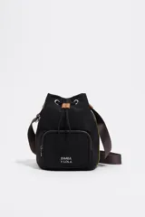 Bolso tipo bucket de Bimba y Lola, confeccionado en nylon negro con detalles en cuero color suela. Cuenta con cierre de cordón ajustable, bolsillo frontal con cierre y correa de hombro regulable de tejido.