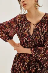 Vestido corto de jacquard con estampado ikat en tonos bordó y detalles metalizados. Presenta mangas largas abullonadas, escote en V con lazo y falda con volados.