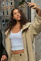 Trench color beige con cuello de solapa subido y manga larga. Tiene bolsillos laterales, forro interior y cinturón de lazo en el mismo tejido. El cuello se cierra con un botón.