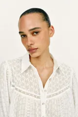 Blusa corta de color blanco con diseño de bordados perforados, cuello solapa, cierre frontal con botones y mangas largas abullonadas.