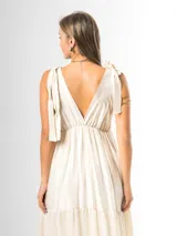 Vestido largo color crudo, confeccionado en mezcla de tencel y lino, con escote en V pronunciado, breteles anchos terminados en lazos en los hombros, y falda larga de varios niveles con volados amplios y fluidos.