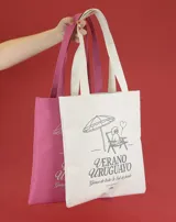 Bolso de mano tipo tote bag color crudo, con asas largas y estampado con motivos veraniegos en azul: sombrilla, reposera, perro caniche y texto "Verano Uruguayo".
