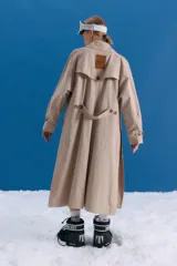 Trench midi de color camel, confeccionado en una mezcla de algodón y nylon con propiedades repelentes al agua. Presenta un diseño clásico con cuello camisero, cierre frontal de botones, bolsillos laterales y tajos en los costados.