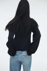Blusa negra de manga larga con corte holgado y tejido texturizado. Presenta cuello corto con volados y abertura frontal con lazos para atar. Tiene canesú con inserciones de encaje y volados sobre los hombros, y puños largos con elástico y volado.