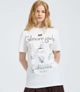 Remera de algodón color blanco con cuello redondo y manga corta. Presenta un estampado temático de la serie Gilmore Girls en la espalda con diseño de taza de café y texto descriptivo.