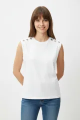 Remera de algodón blanca, de corte holgado y sin mangas, con cuello a la base y detalle de ojalillos metálicos sobre los hombros.