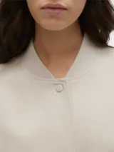 Campera bomber corta de color beige, confeccionada en tejido sarga, con cuello tipo rib, cierre frontal de botones a presión, bolsillos frontales con solapa y puños y dobladillo elasticados.