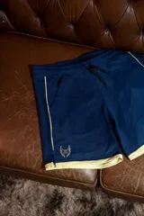 Short deportivo de lycra color azul marino con cordón de ajuste negro. Presenta ribetes y un pequeño logo de alas en color dorado/beige en la parte inferior izquierda.