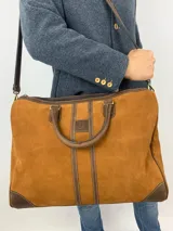 Bolso de cuero color tostado con acabado de gamuza y detalles en cuero liso marrón oscuro. Presenta un diseño con dos asas de mano, una correa larga ajustable y removible para el hombro, y cierre superior de cremallera. Incluye franjas verticales decorativas en el centro con el logo de la marca grabado.
