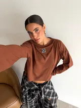 Remera de manga larga color marrón, con cuello redondo y detalle de parche cuadrado con logo bordado en el centro del pecho. Presenta un corte holgado y diseño minimalista.