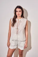 Blusa sin mangas de algodón color blanco off white, con cuello redondo y cierre frontal mediante cordones para atar. Presenta delicados detalles de bordados tonales e inserciones de encaje vertical en el frente.
