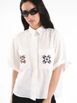 Camisa de manga corta color blanco, con calce oversized, hombros caídos y cierre frontal con botones ocultos. Presenta bolsillos delanteros con bordados geométricos y mangas con dobladillo.