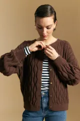 Cardigan de punto color terracota con diseño de trenzas verticales, cuello redondo y cierre frontal con botones metálicos. Presenta mangas largas con puños ajustados y un calce holgado.
