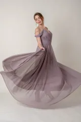 Vestido largo de fiesta color lila, confeccionado en gasa. Presenta un corsé drapeado con escote off-shoulder y mangas caídas. La falda es amplia y fluida.