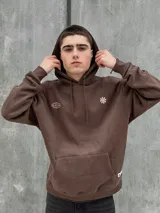 Canguro marrón con capucha ajustable con cordones, bolsillo tipo canguro y puños elásticos. Presenta un logo de un globo terráqueo y una flor bordados en el pecho.