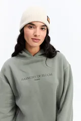 Canguro de algodón color beige con capucha amplia y corte relajado. Presenta un estampado rectangular en la espalda con motivos florales en tonos azules y el texto 'Harmony of Bloom'.