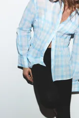 Camisa de manga larga con estampado de cuadros vichy en tonos celeste y blanco. Presenta cuello clásico, cierre frontal con botones y detalle de fruncido en la parte central delantera.