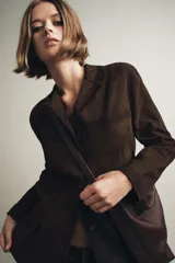 Blazer de mujer color marrón oscuro, confeccionado en piel de ante 100%. Presenta cuello solapa de muescas, manga larga, bolsillos de plastrón frontales y cierre frontal de botones.