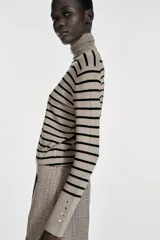Jersey de punto liso en color beige, con cuello alto y manga larga con detalle de botones metálicos en los puños.