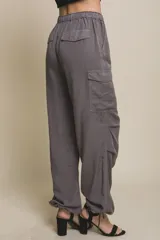 Pantalón cargo gris de corte ancho y relajado, confeccionado en tela fluida. Cuenta con cintura elástica con cordón de ajuste, bolsillos laterales, bolsillos cargo en las piernas y detalles de fruncido con cordones ajustables en los tobillos.