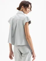 Camisa de cuero ovino color gris claro, con cuello camisero, volados centrales y mangas cortas con volumen generado por frunces en la copa. Cierre frontal con broches forrados.